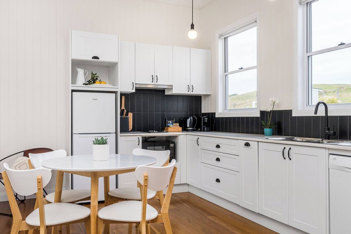 A modern kitchen with white cabinets, a black backsplash, and a wooden dining table with chairs.