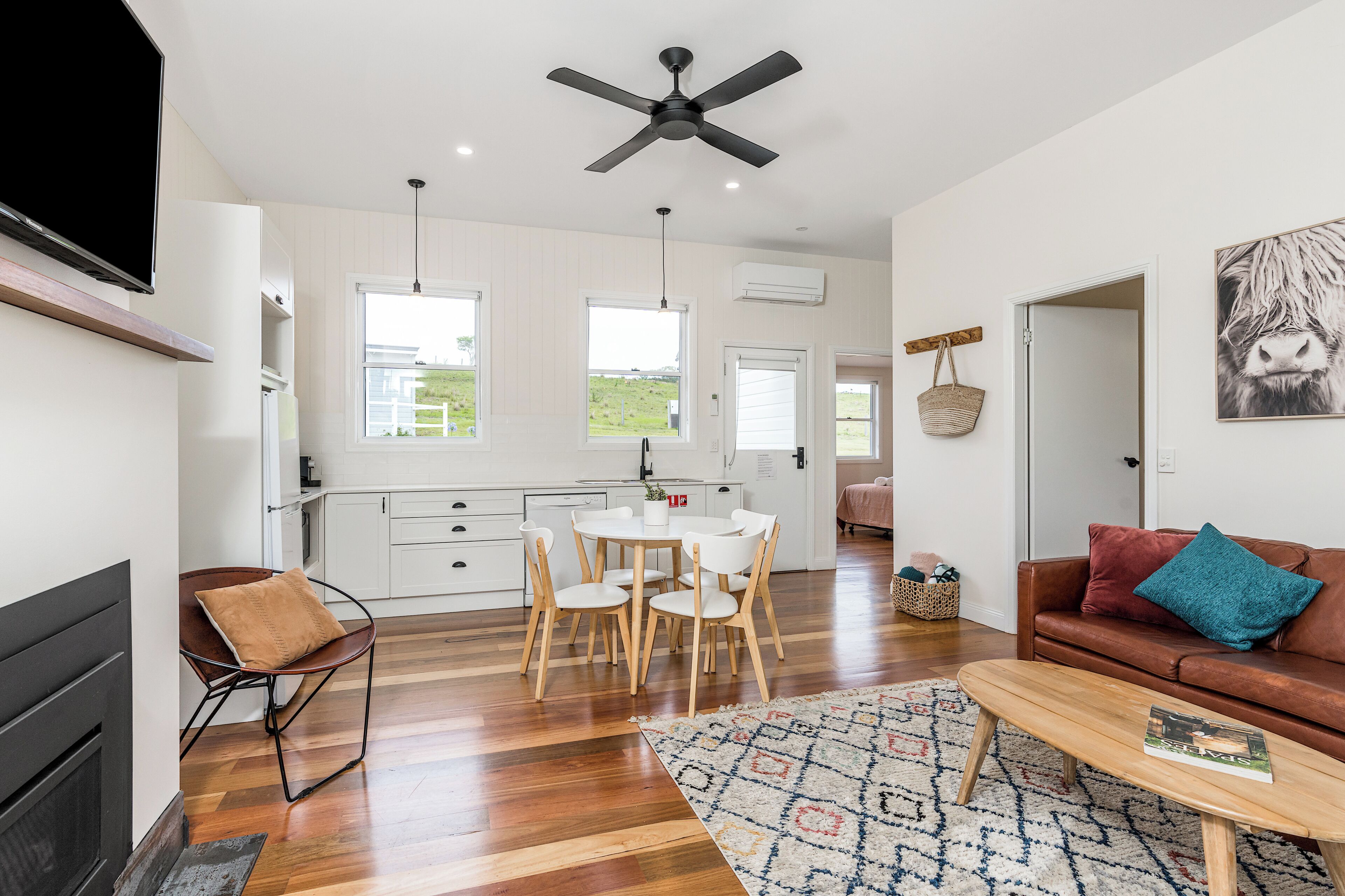 A modern kitchen with white cabinets, a dining area with a round table and chairs, a brown leather sofa with a teal pillow, and a fireplace.