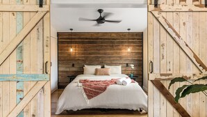 Une chambre avec une tête de lit en bois, un ventilateur de plafond, un lit recouvert d’une couverture à rayures et deux lampes suspendues.