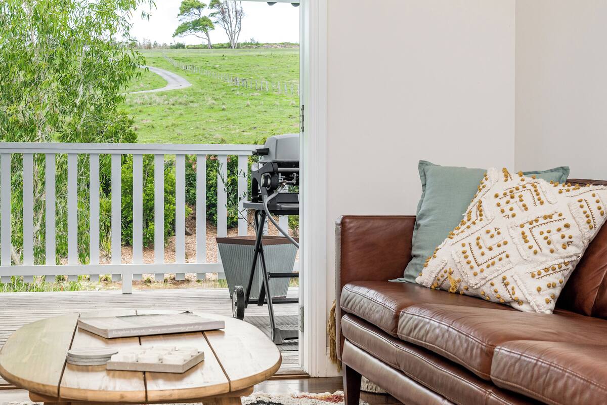 A room with a brown leather sofa, a wooden coffee table with books, a white railing, and a view of a green field.