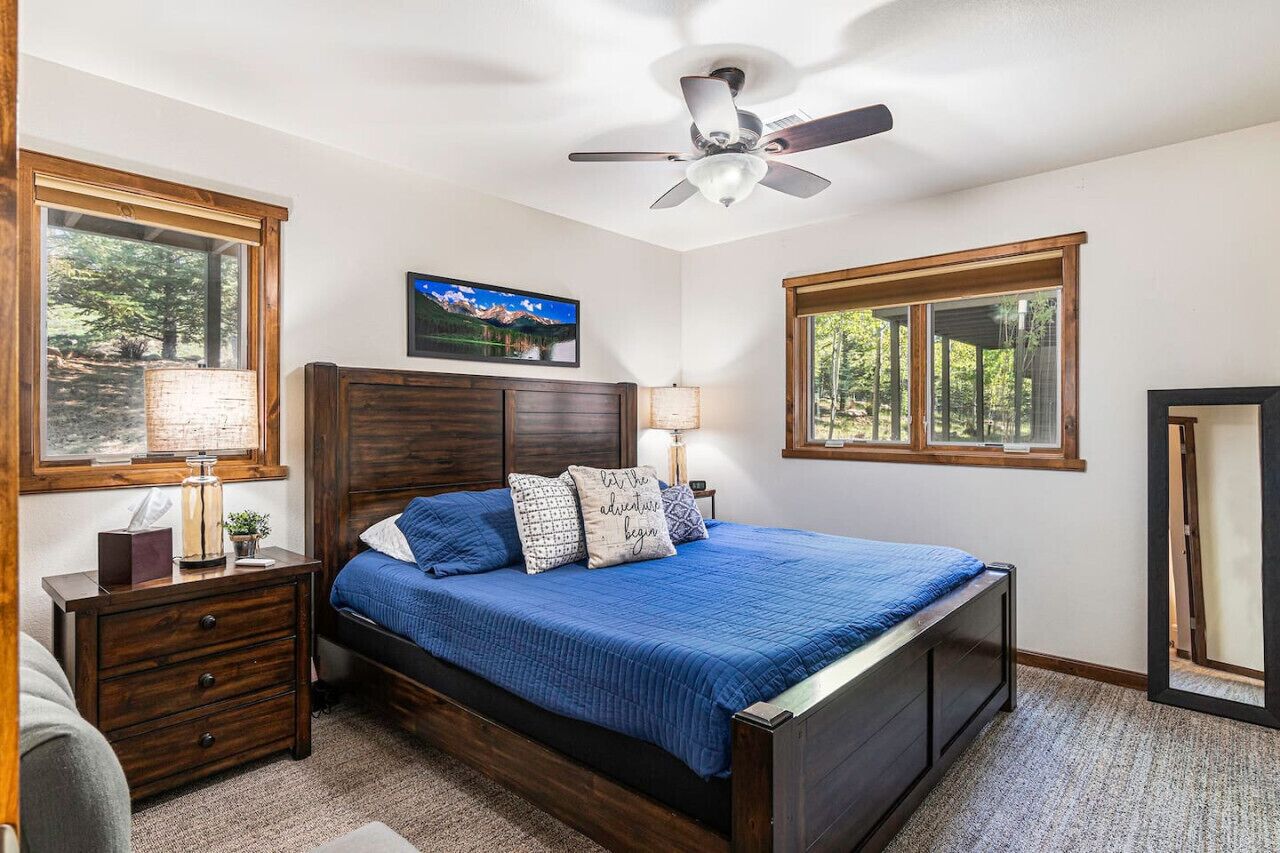 Un dormitorio con cama, cabecera de madera, ventilador de techo, mesita de noche, lámpara y ventana con vista a árboles.