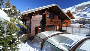 Una cabaña de madera con balcón, rodeada de árboles nevados y una montaña al fondo.