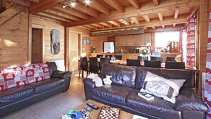 Una cabaña de madera acogedora, con una sala de estar que incluye un sofá de cuero, una mesa de centro con un juego de ajedrez y una zona de cocina con armarios de madera y una mesa de comedor.