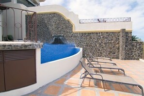 A pool area with a stone wall, a fountain, and lounge chairs.