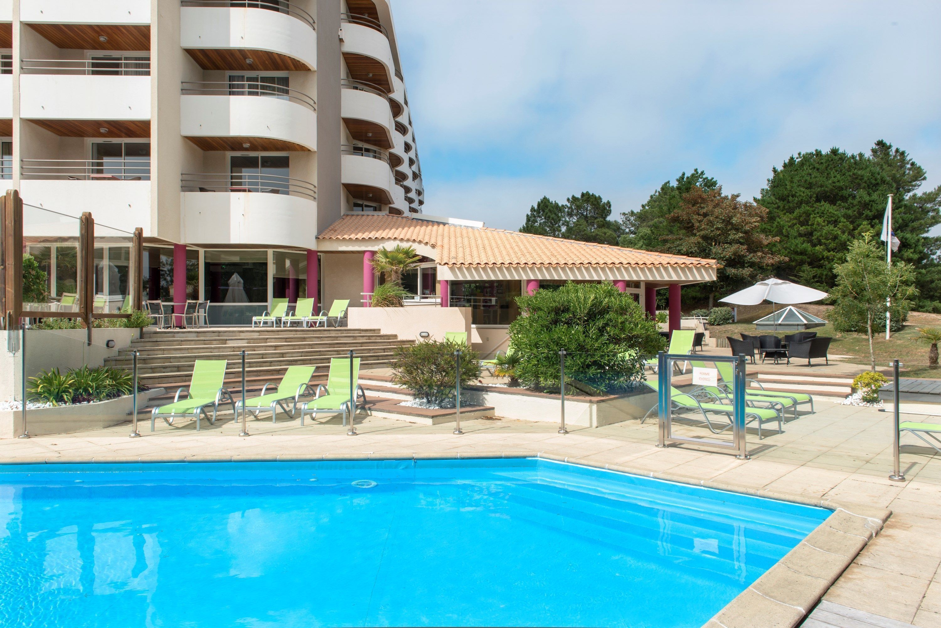 Une piscine avec des chaises longues, un espace de détente couvert et un bâtiment doté de balcons.