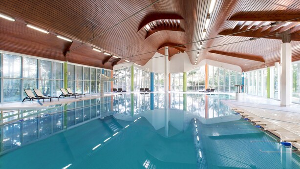 En stor indendørs swimmingpool med trælofter, glasvægge og liggestole langs kanten.