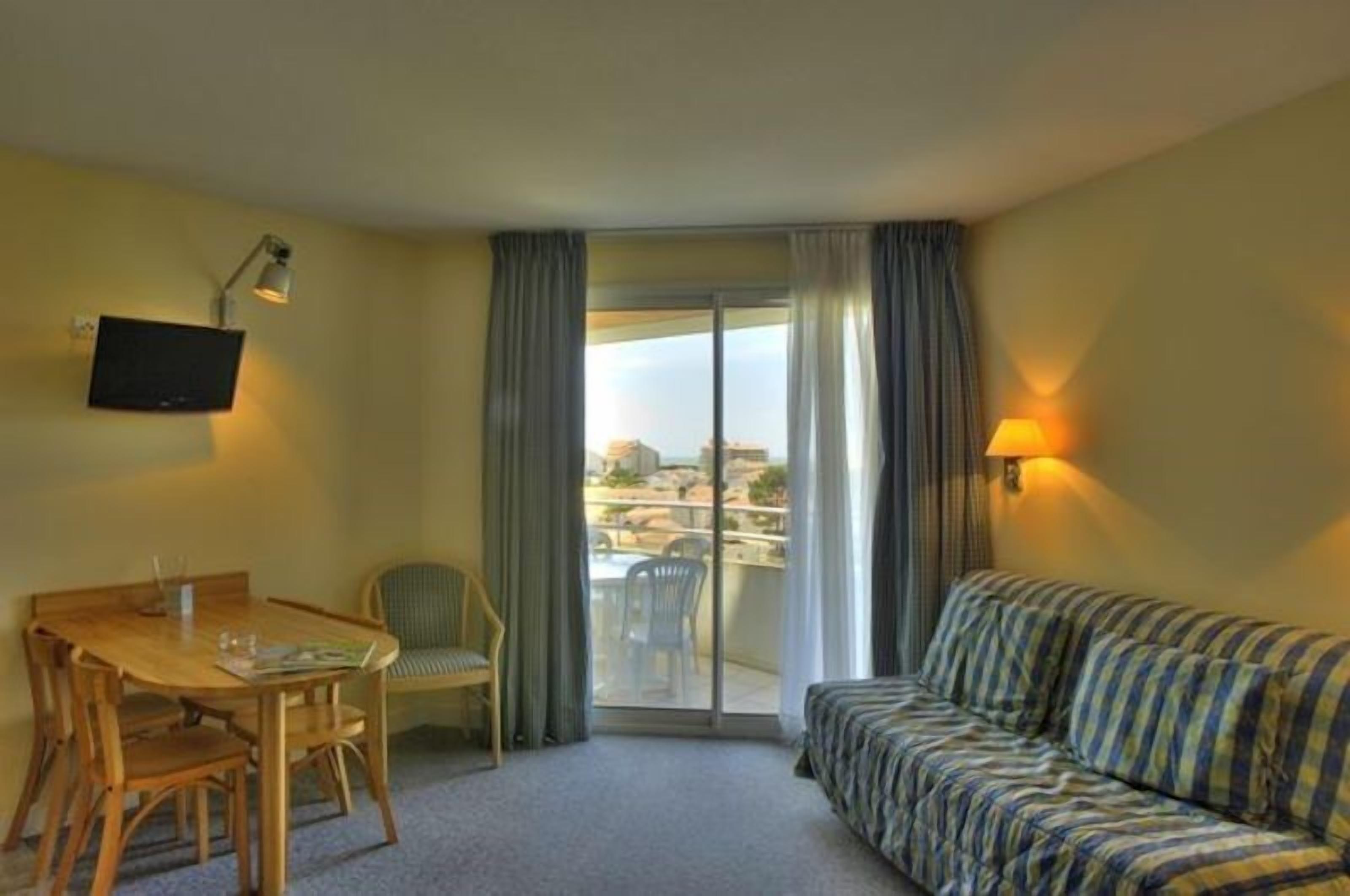 A hotel room with a wooden dining table, a sofa, a TV mounted on the wall, and a balcony with a view of the city.
