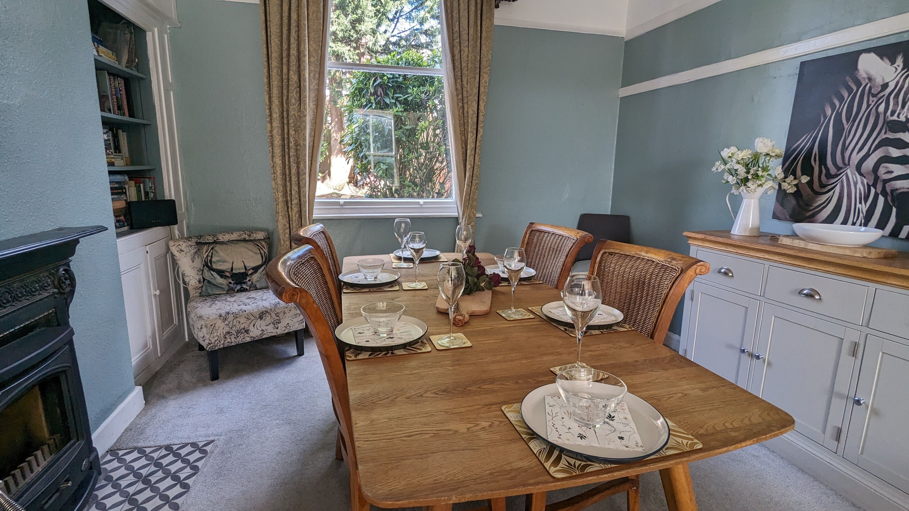 A dining room with a wooden table set for six, flanked by wicker chairs, a fireplace, and a bookshelf.