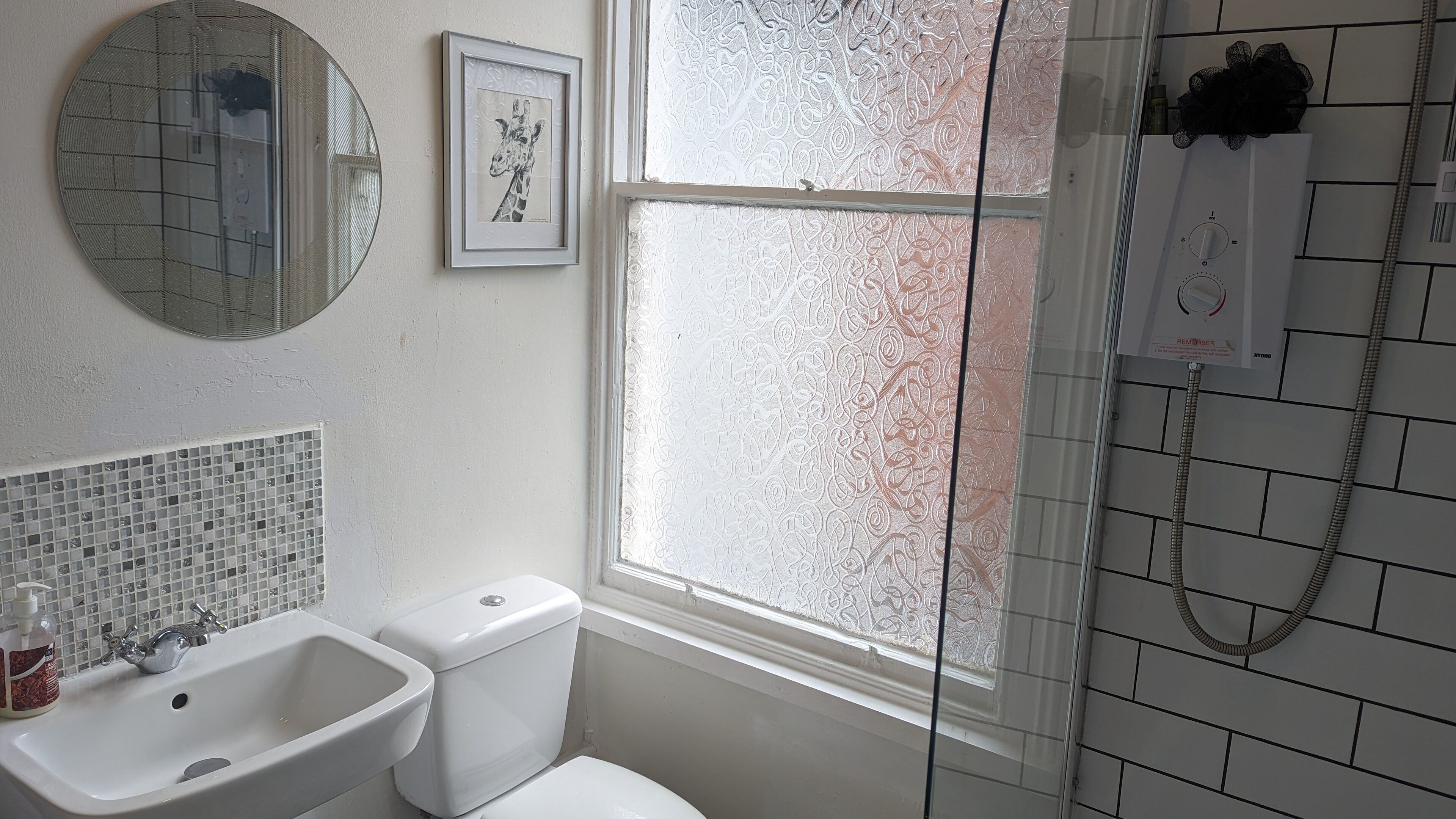 A bathroom with a toilet, sink, and shower. There is a window with frosted glass, a mirror, and a framed picture on the wall.