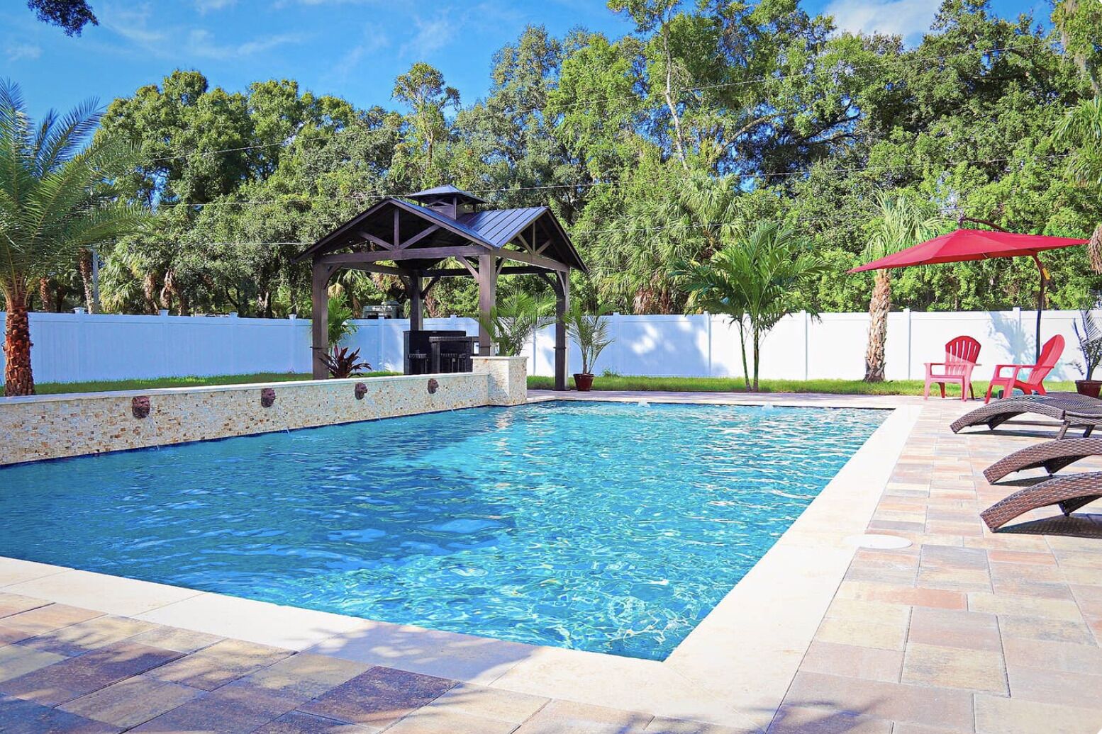 Una piscina rectangular con solárium, un pabellón y sillas rojas.