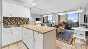 A modern kitchen with white cabinets, a granite countertop, and a large island. There is a living area with a sofa, a coffee table, and a TV. The room has large windows and a view of the outside. - Steps to Private Beach! Heated pool-tennis-picklebal-shuffleboard. (Destin)