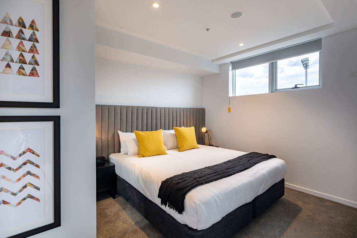 A modern bedroom with a large bed, a grey headboard, and a window with a view of a tall structure.
