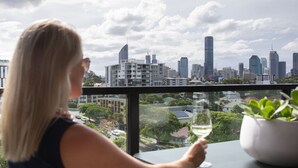 Una persona sosteniendo una copa de vino, contemplando un paisaje urbano con edificios modernos y una vista de la ciudad a través de un ventanal.