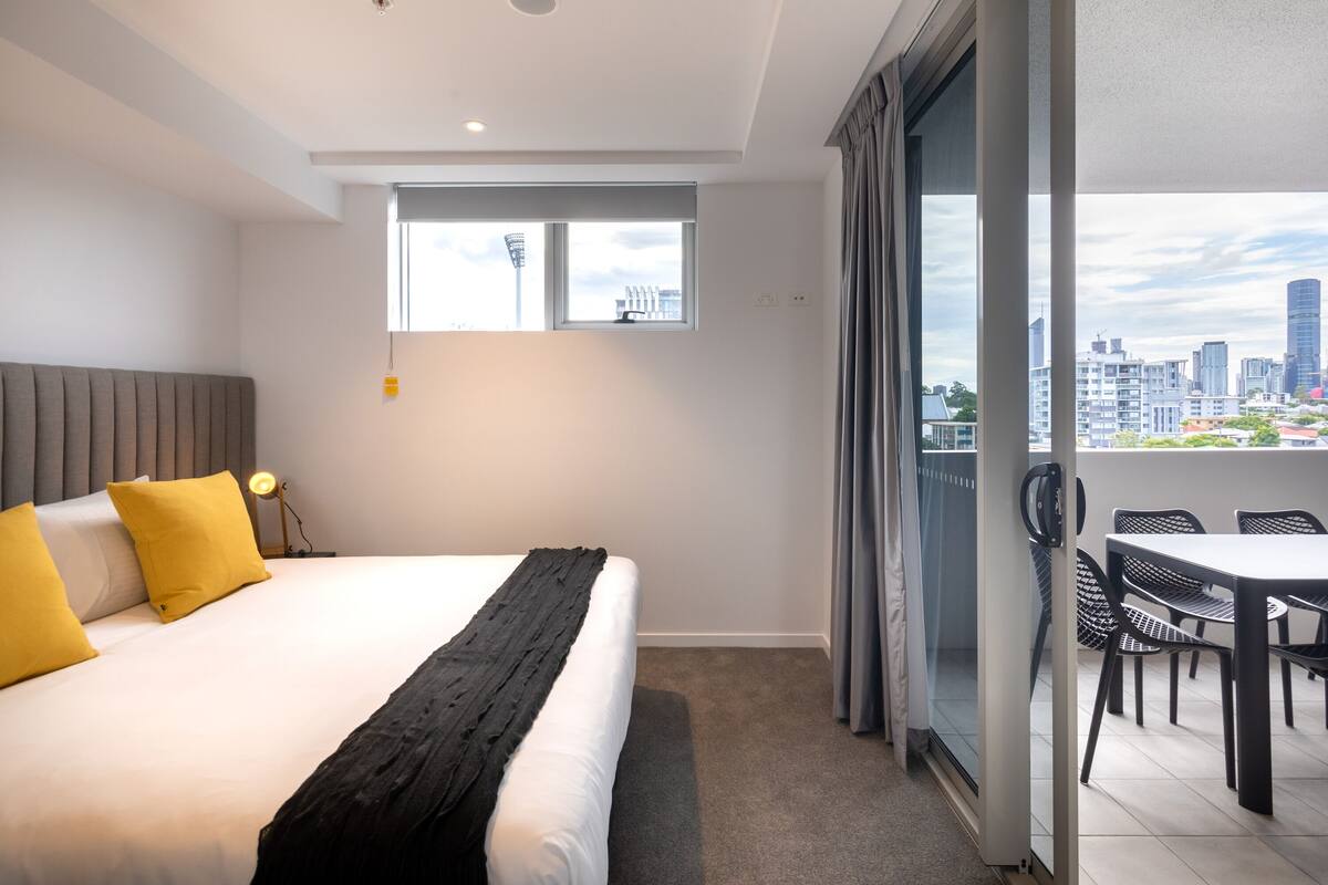 A hotel room with a bed, a nightstand, a window with a city view, and a balcony with a table and chairs.