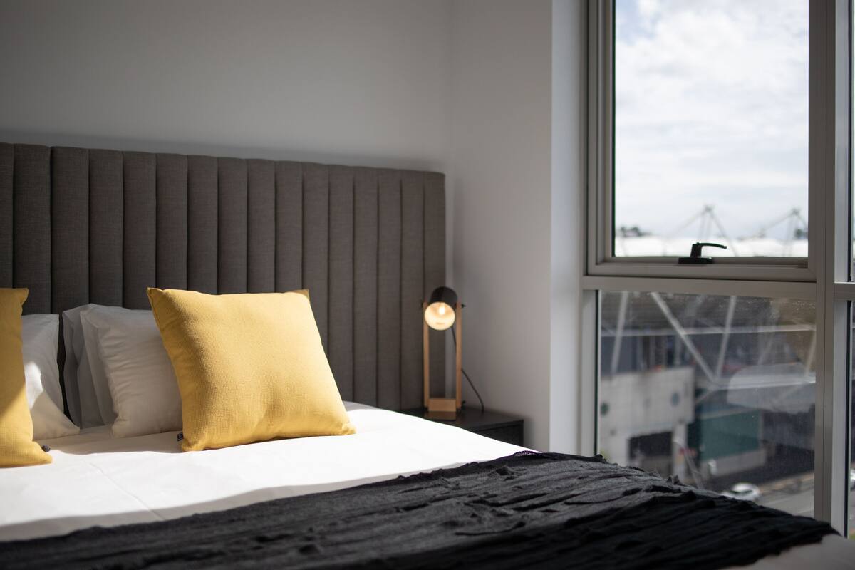 A modern bedroom with a bed, pillows, a bedside lamp, and a window with a view of buildings.