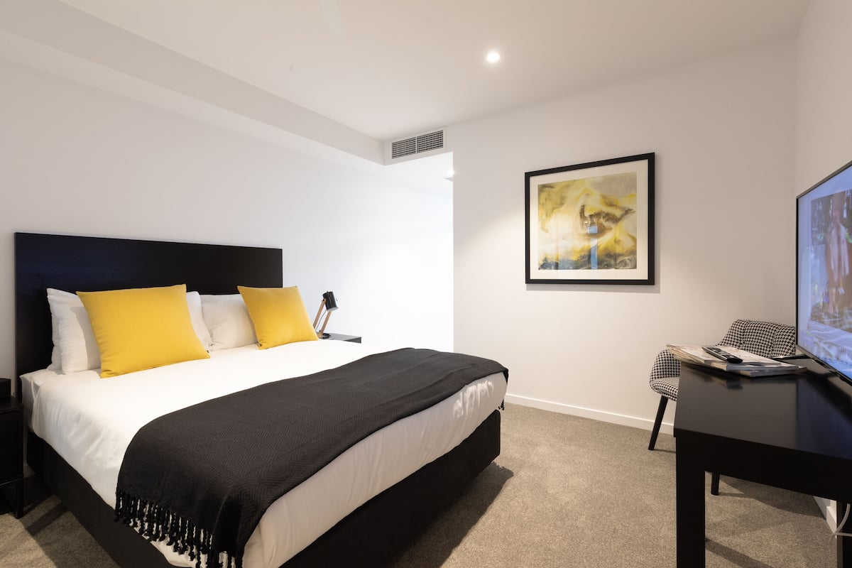 A modern bedroom with a large bed, a desk, and a framed artwork on the wall.