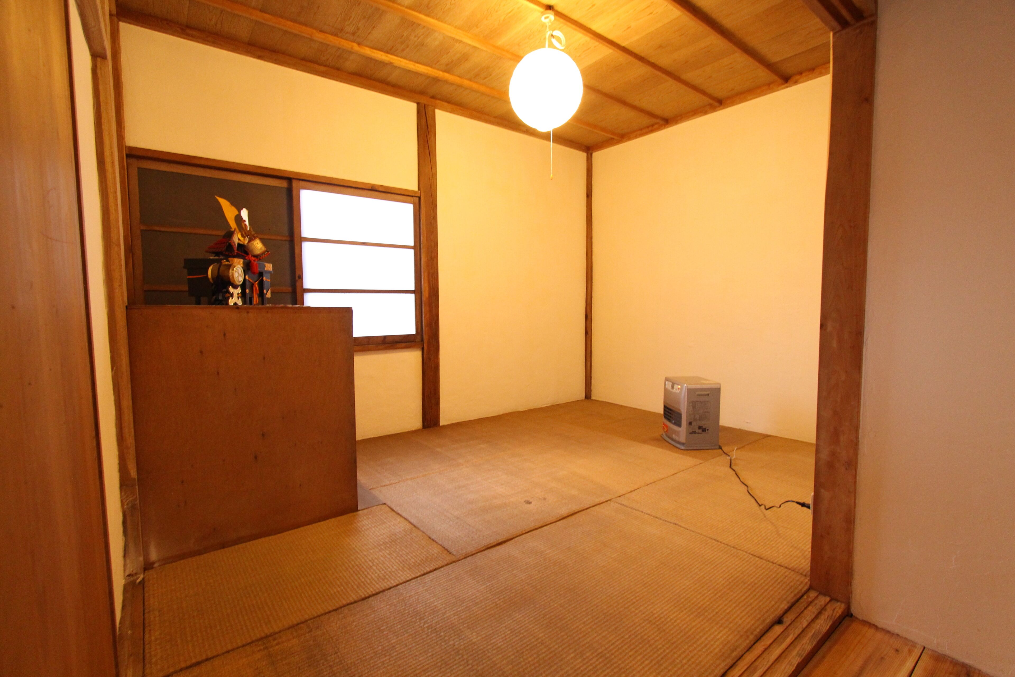 A room with tatami flooring, a wooden desk with a decorative item, a standing fan, and a hanging light fixture.