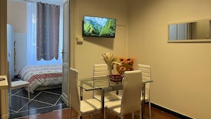 A hotel room with a bed, a TV mounted on the wall, a glass dining table with chairs, and a vase with flowers. - Appartamento a Invrea Posto Auto Incluso (Genoa)
