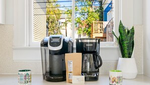 A coffee maker, a coffee bag, and two mugs on a kitchen counter by a window. - Elegance Meets Ease in Historic CHS | The Rosewood (Charleston)