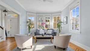 A spacious living room with a sofa, two armchairs, a coffee table, and large windows offering a view of the outside. - Elegance Meets Ease in Historic CHS | The Rosewood (Charleston)
