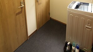 A laundry room with two wooden doors, a window with blinds, and a washing machine with bottles of detergent. - Gran Kodachi (Fujikawaguchiko)