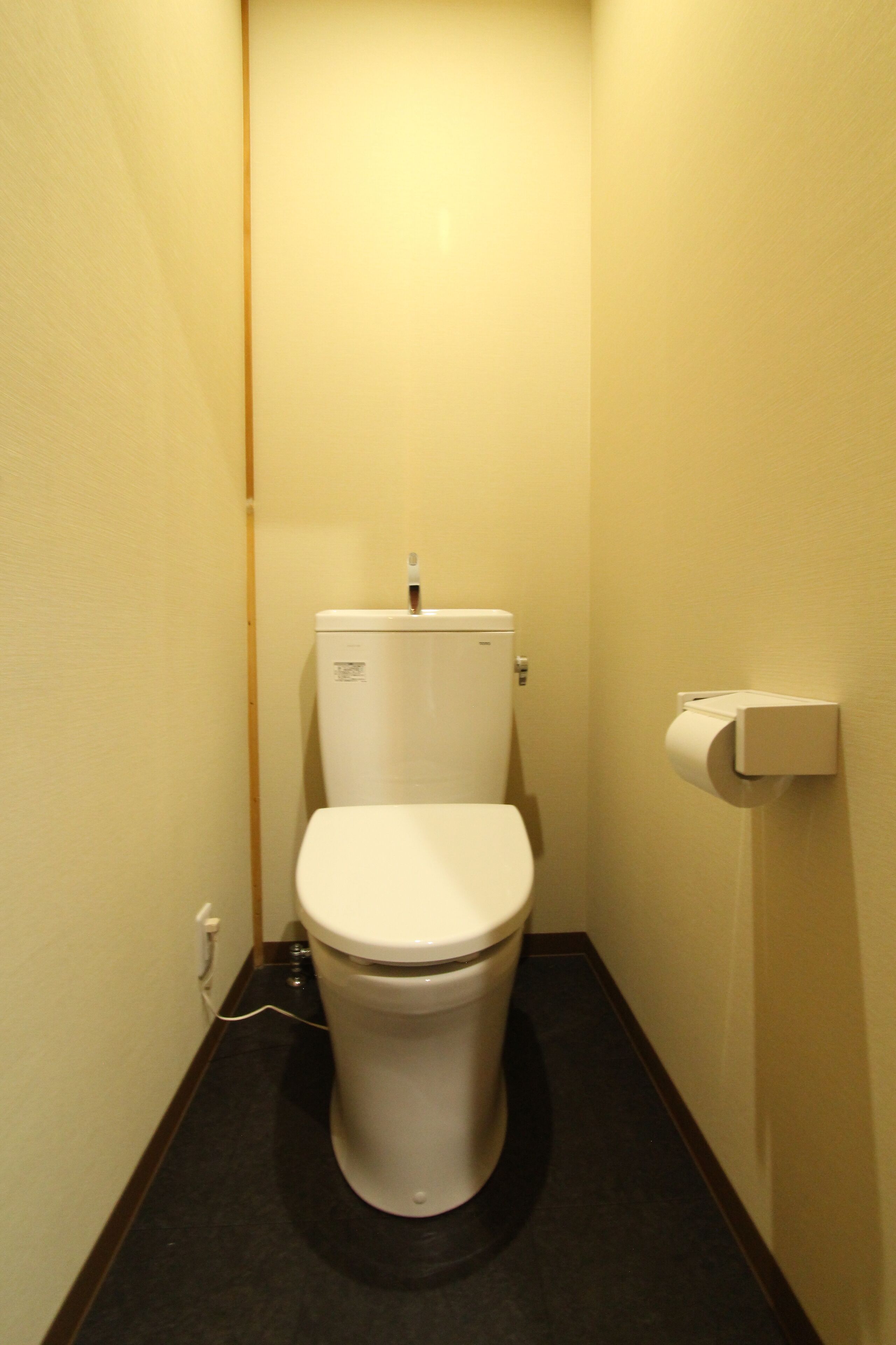 A small, narrow bathroom with a white toilet, a paper towel dispenser, and a visible electrical outlet on the wall.