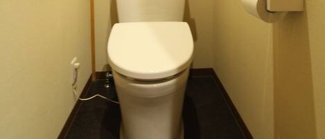A small, narrow bathroom with a white toilet, a paper towel dispenser, and a visible electrical outlet on the wall.
