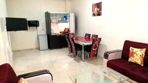 Un ambiente compatto con divano, tavolino in vetro, tavolo da pranzo con sedie, angolo cottura e televisore piatto a parete.
