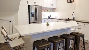 Une cuisine moderne avec des meubles blancs, un réfrigérateur en acier inoxydable, un grand îlot central doté de tabourets et un luminaire suspendu.