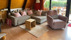 A cozy living room with a sofa, armchair, and a small wooden table. There is a red curtain, a lamp, and a view of the outdoors through a glass door.