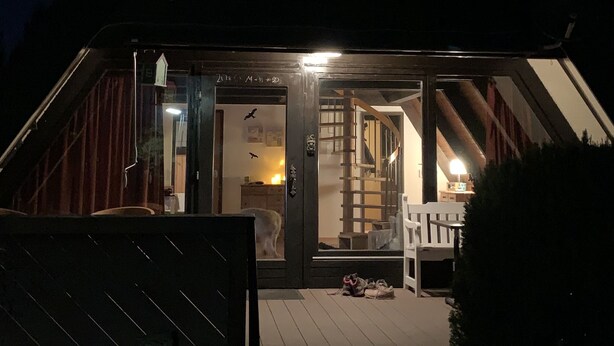 A wooden cabin with a porch, a staircase leading to the entrance, and a lit interior visible through the glass doors.
