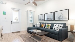A modern living room with a grey sofa, a coffee table, and nautical-themed pillows. There are framed pictures on the wall and a ceiling fan. - Sunny Margaritaville Cottage - Fully Equipped Kitchen, Welcoming Living Area (Kissimmee)