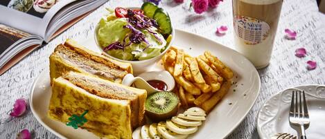 Un plato de comida que incluye tostadas, papas fritas, una ensalada y kiwi en rodajas, con una bebida y un libro al fondo.