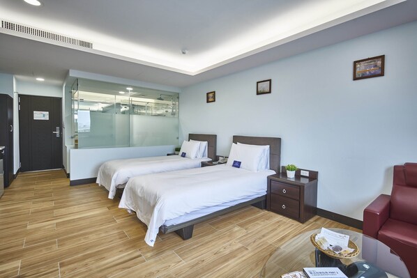 Habitación de hotel con dos camas, un escritorio, una silla, una mesita con una cesta y una pared divisoria de vidrio.
