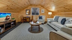 A cozy attic room with wooden walls and ceiling, a TV, a sofa, a coffee table, and a dining table with chairs. - SHADE MOUNTAIN RETREAT ★ CEDAR BARREL SAUNA & HOT TUB!! ★ 28 ACRES ★ SLEEPS 20 (East Waterford)