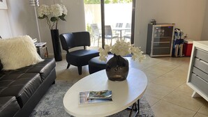 A living room with a black leather sofa, a white coffee table with a magazine, a vase with white flowers, and a view of the outside patio.