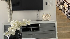 A modern living room with a flat-screen TV mounted on the wall, a grey entertainment unit below, a round white table with a magazine, a black vase with white flowers, and a decorative vase with peacock feathers.