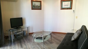 A living room with a flat-screen TV on a glass table, a dark sofa with a patterned pillow, and two framed artworks on the walls. - 1 Bedroom Apt - 2 Nt Min(Comfortable one bedroom apartment with city view) (Spring Hill)