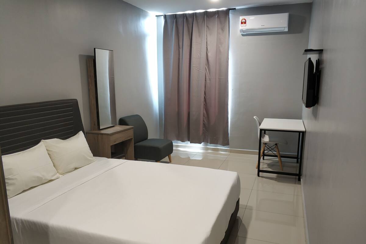 A hotel room with a bed, a desk, a chair, a mirror, and an air conditioning unit.