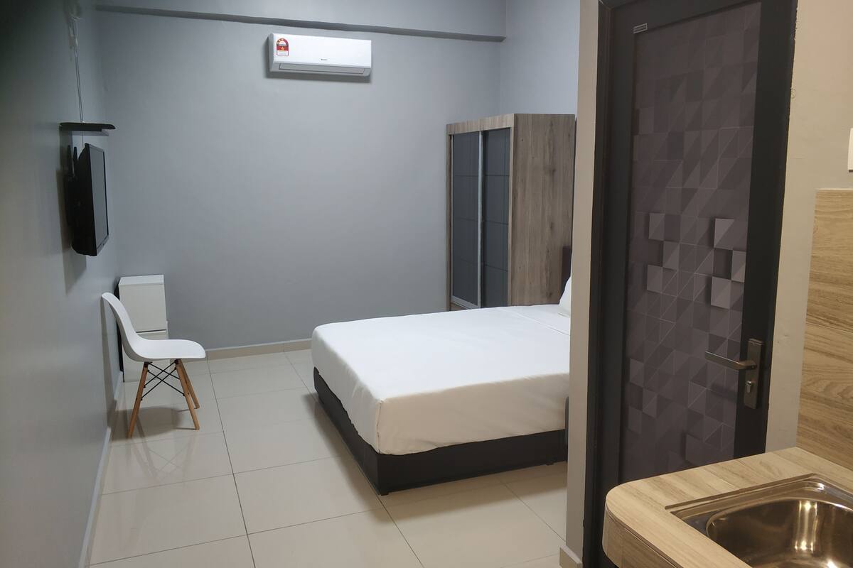A modern hotel room with a bed, a desk, a chair, a wardrobe, and a bathroom with a sink and mirror.