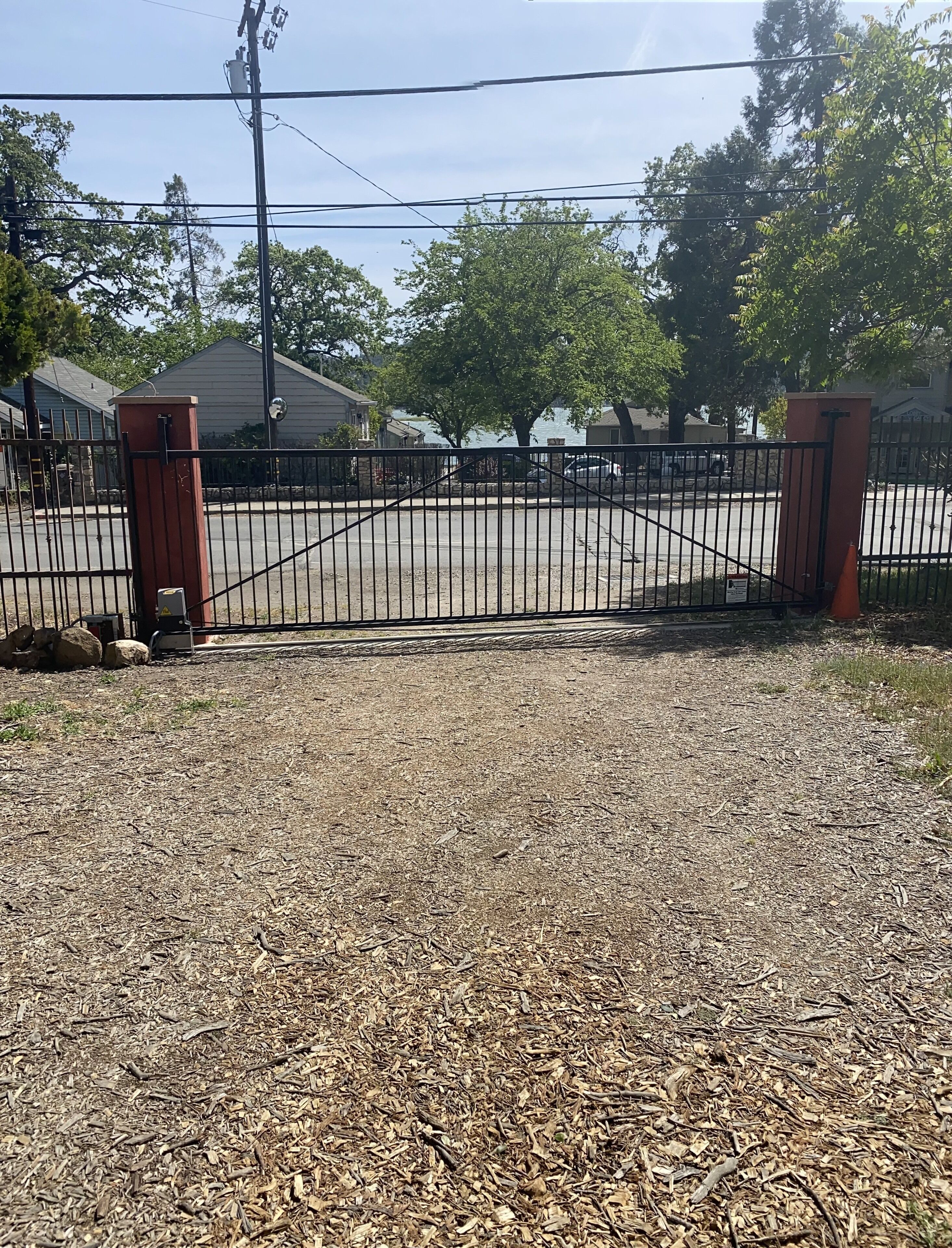 A gated parking area with a gravel surface, surrounded by trees and a building with a sloped roof.
