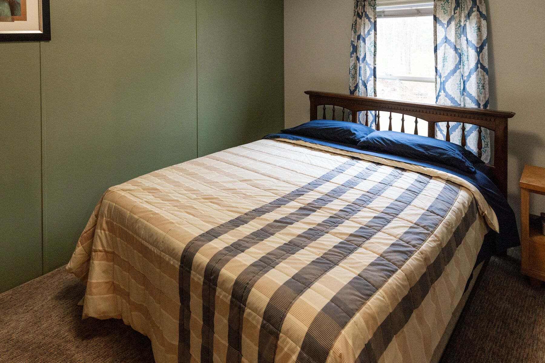 Un lit simple avec une couvre-lit à rayures, une tête de lit en bois et une fenêtre ornée de rideaux à motifs.
