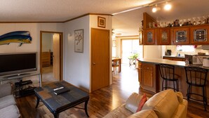 A living room with a TV, a sofa, and a coffee table. There is a kitchen area with wooden cabinets and a countertop. - Motorsports Basecamp near lakes and forest (Hayward)