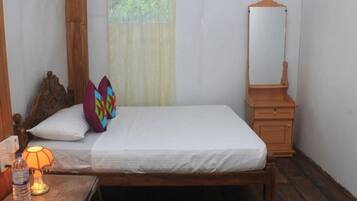 A neatly made bed with a white sheet, a wooden headboard, and colorful pillows. There is a small bedside table with a lamp and a bottle of water. A wooden nightstand with a mirror is to the right of the bed.