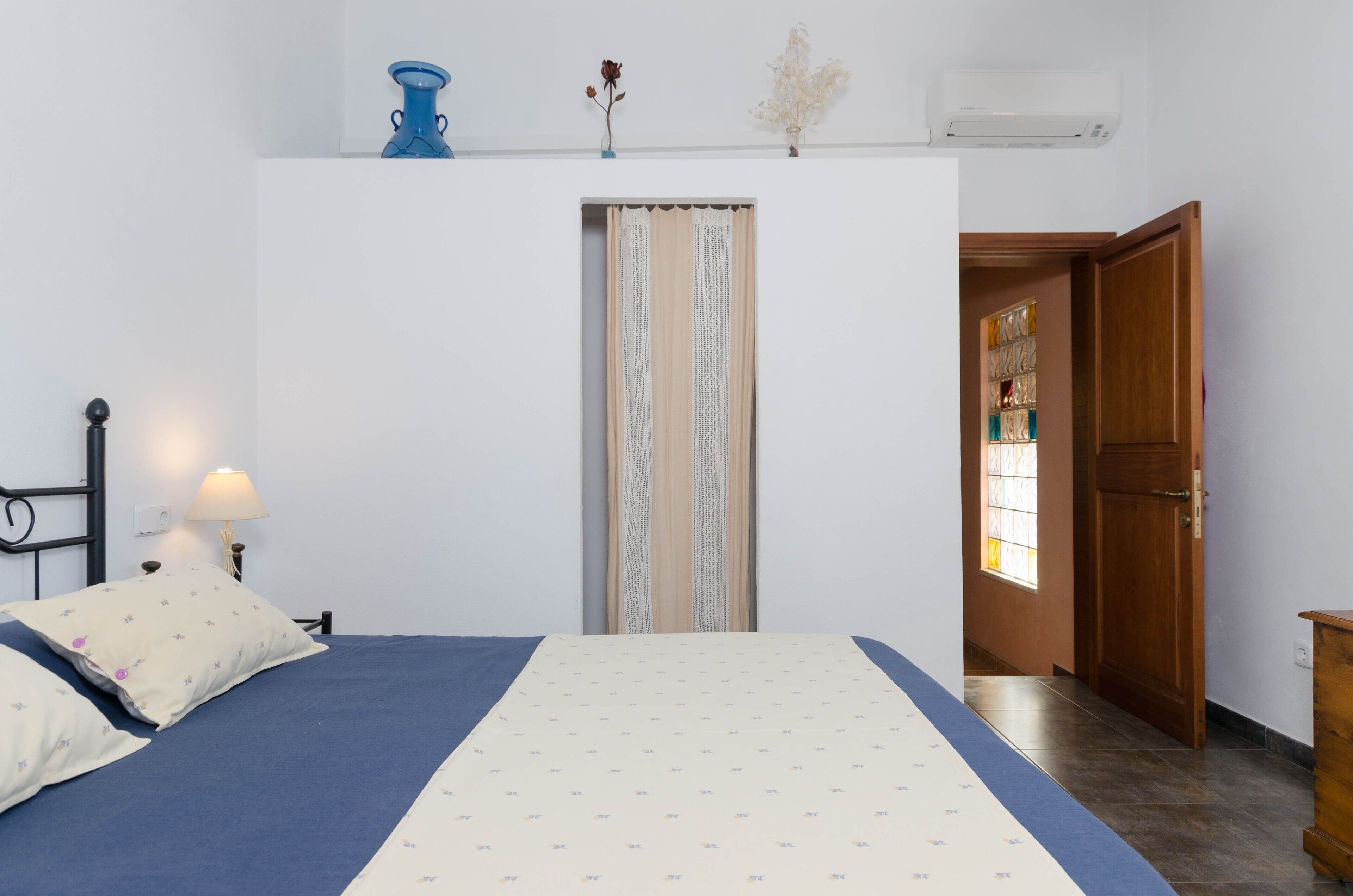A bedroom with a bed, a nightstand, a lamp, a window with curtains, and a door leading to another room.