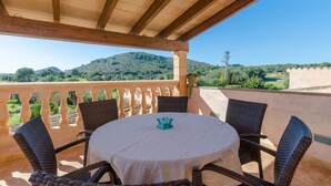 Un espacio de estar al aire libre cubierto, con sillas de mimbre y una mesa, con vistas a un paisaje pintoresco con colinas y cielo despejado.