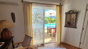 Una habitación con piso de madera, alfombra blanca, una silla, una mesa con un jarrón, un espejo en la pared y vistas a una piscina y montañas a través de una puerta.