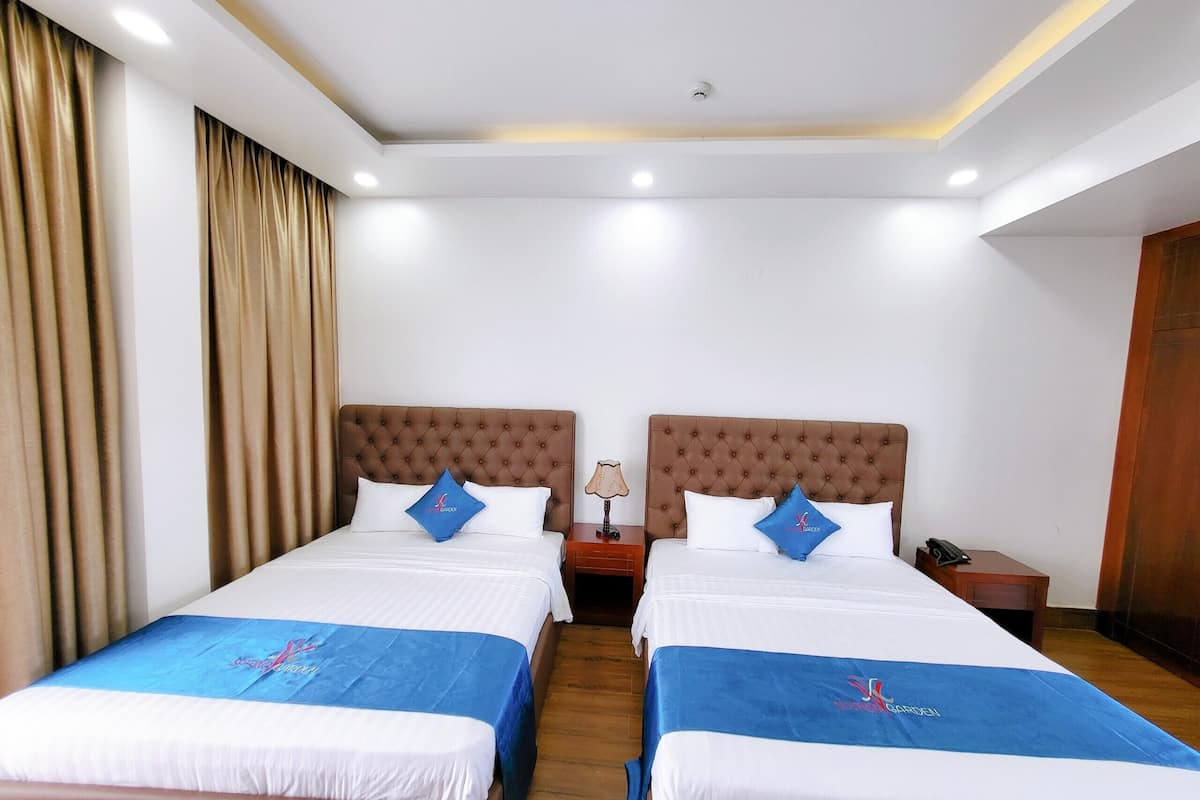 A hotel room with two beds, each with a blue bedspread and matching pillows, a wooden headboard, and a nightstand with a lamp.