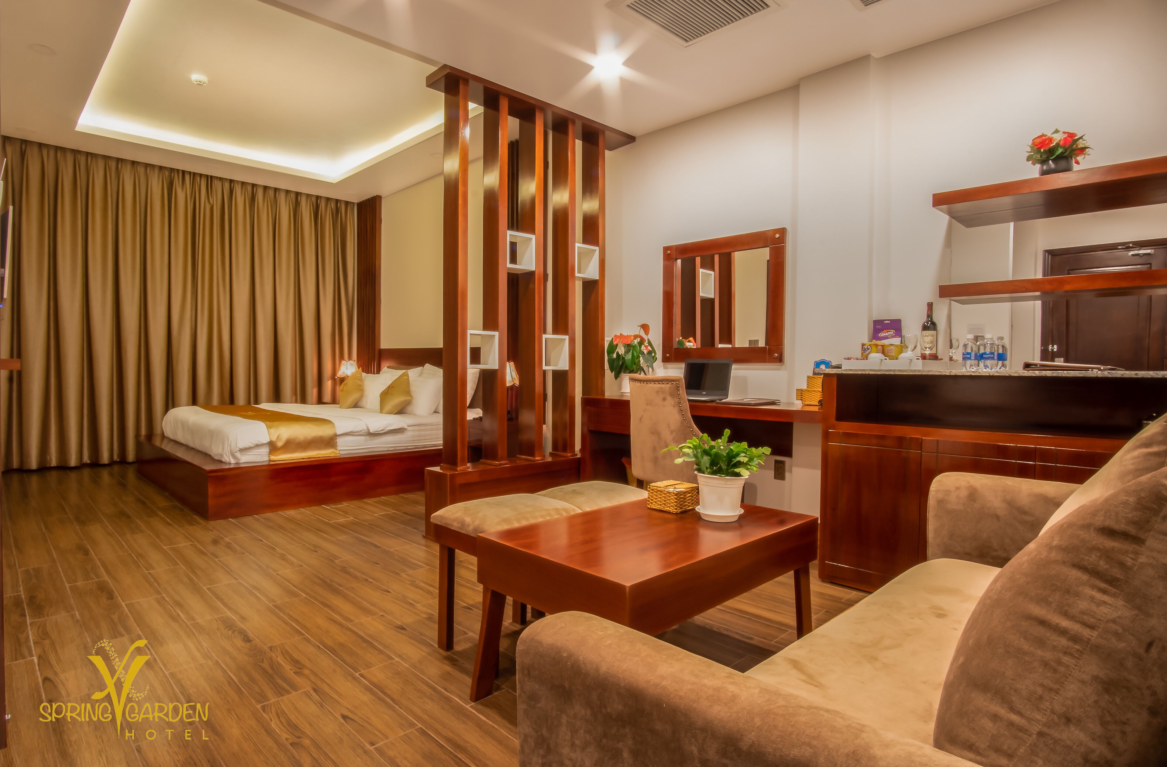 A hotel room with a bed, a desk, a chair, a sofa, a potted plant, and a mirror.
