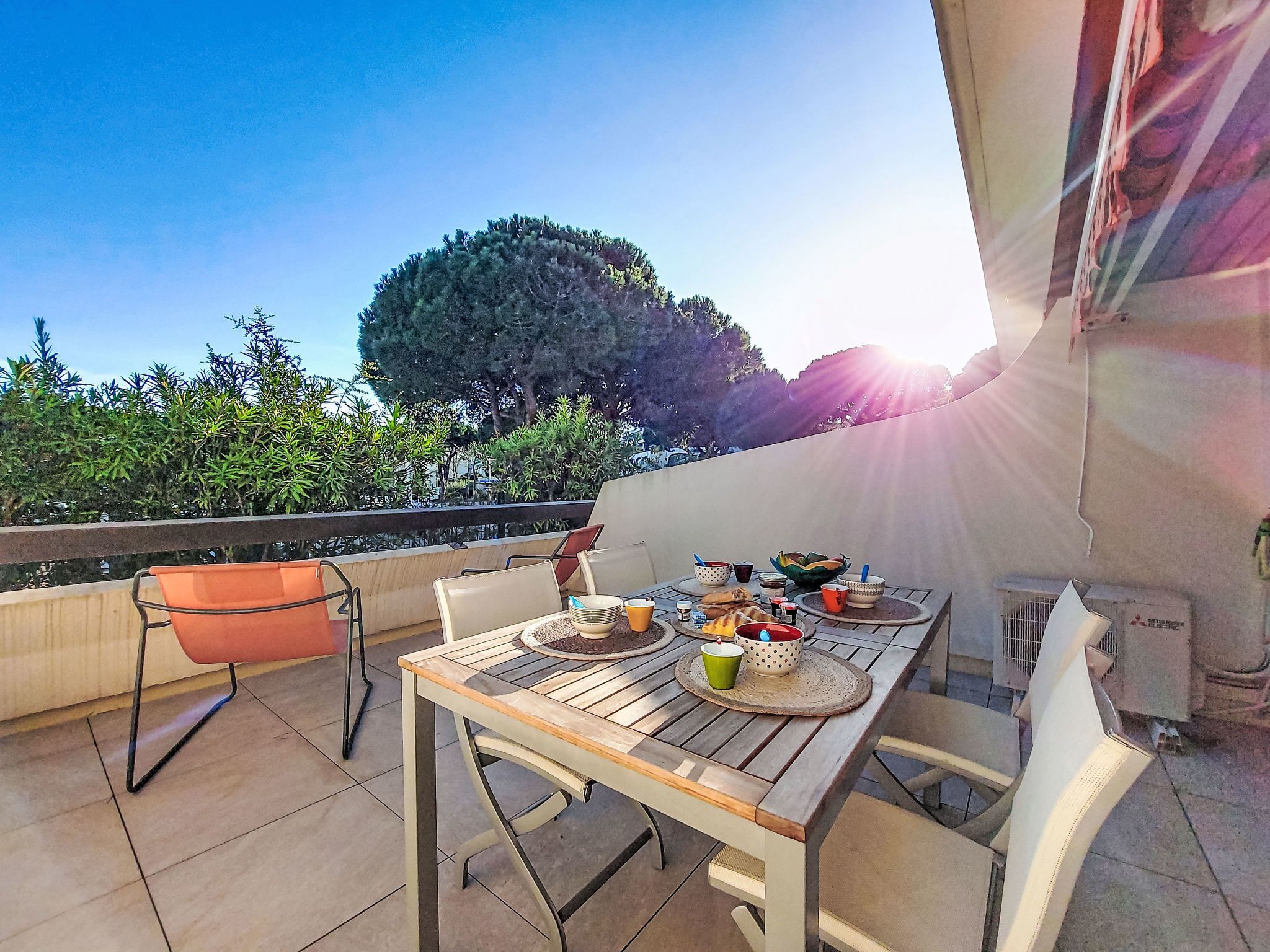 Ein Essbereich im Freien mit einem Tisch für zwei Personen, umgeben von Stühlen, auf einer Terrasse mit Blick auf einen großen Baum und einen klaren Himmel.